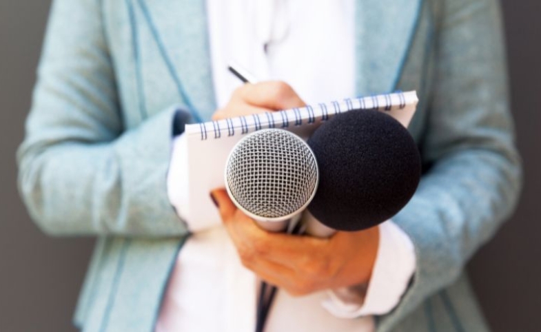 A woman writing on a notepad and holding two microphones by Mihajlo Maricic via Canva.com