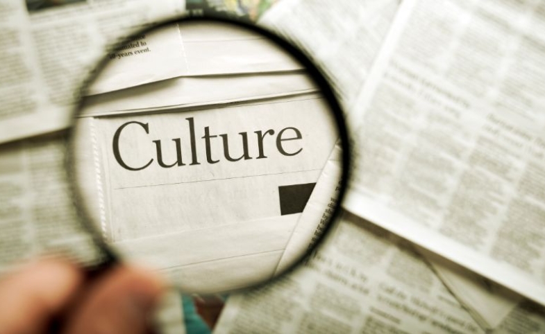 A hand holding a magnifying glass over a newspaper that has the word "culture" by fotosipsak via Canva