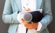 A woman writing on a notepad and holding two microphones by Mihajlo Maricic via Canva.com